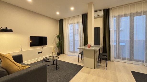 Una sala de estar moderna con un sofá, una mesita de centro, un televisor montado en la pared y un balcón con cortinas.