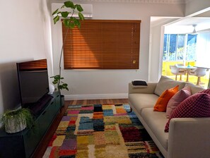 A living room with a sofa, a television, a colorful rug, a window with blinds, and a plant. - Super cute house that's full of character. Close to airport and train station. (Wynnum)