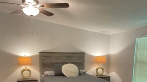 A bedroom with a bed, wooden headboard, two bedside tables with lamps, a ceiling fan, and a window with blinds. - Charming 3-bedroom 2 Bath farmhouse on 7.4 Acres (Trenton)