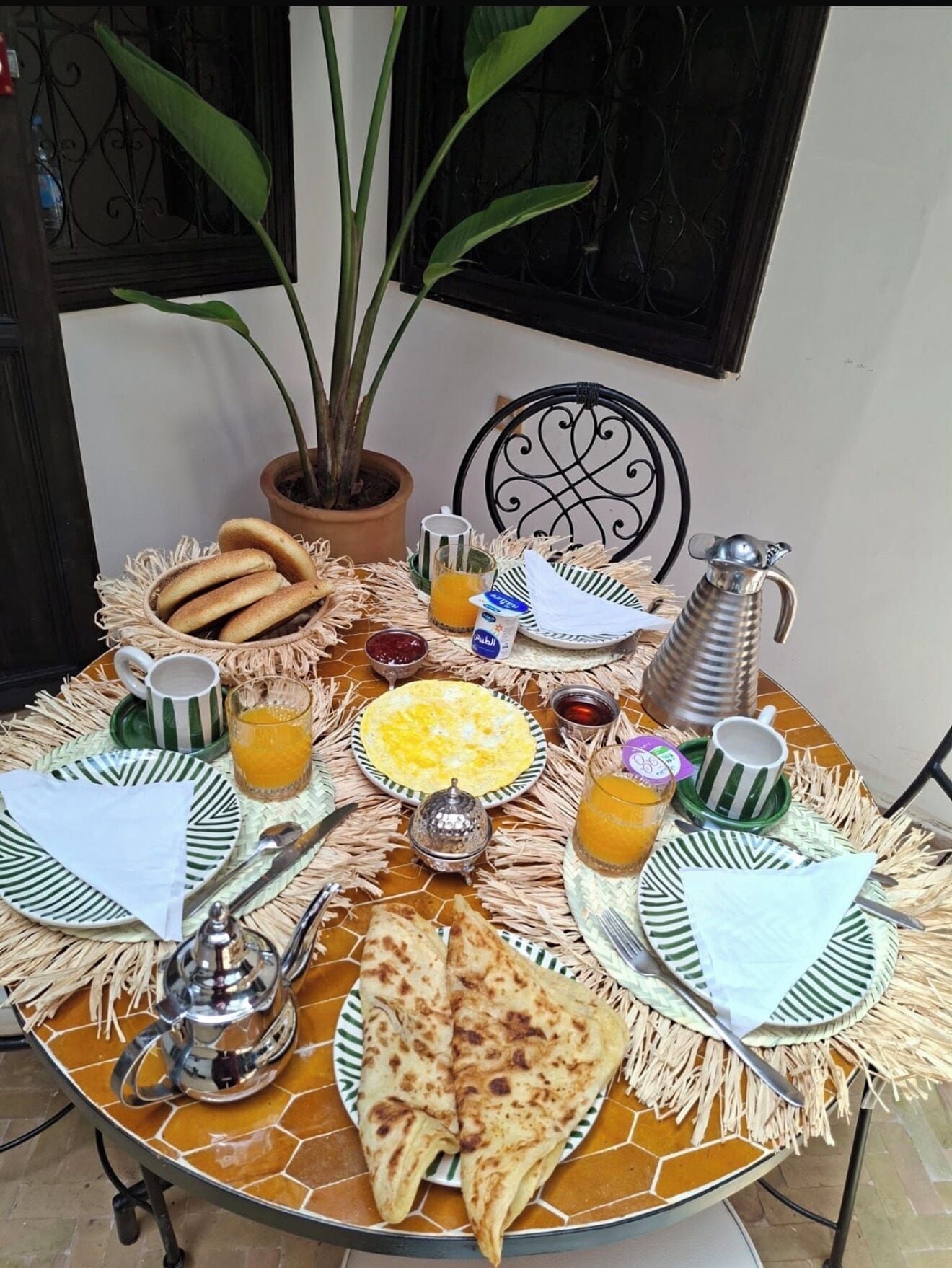 A table set with breakfast items including bread, eggs, and beverages, with a potted plant and a decorative metal chair in the background.