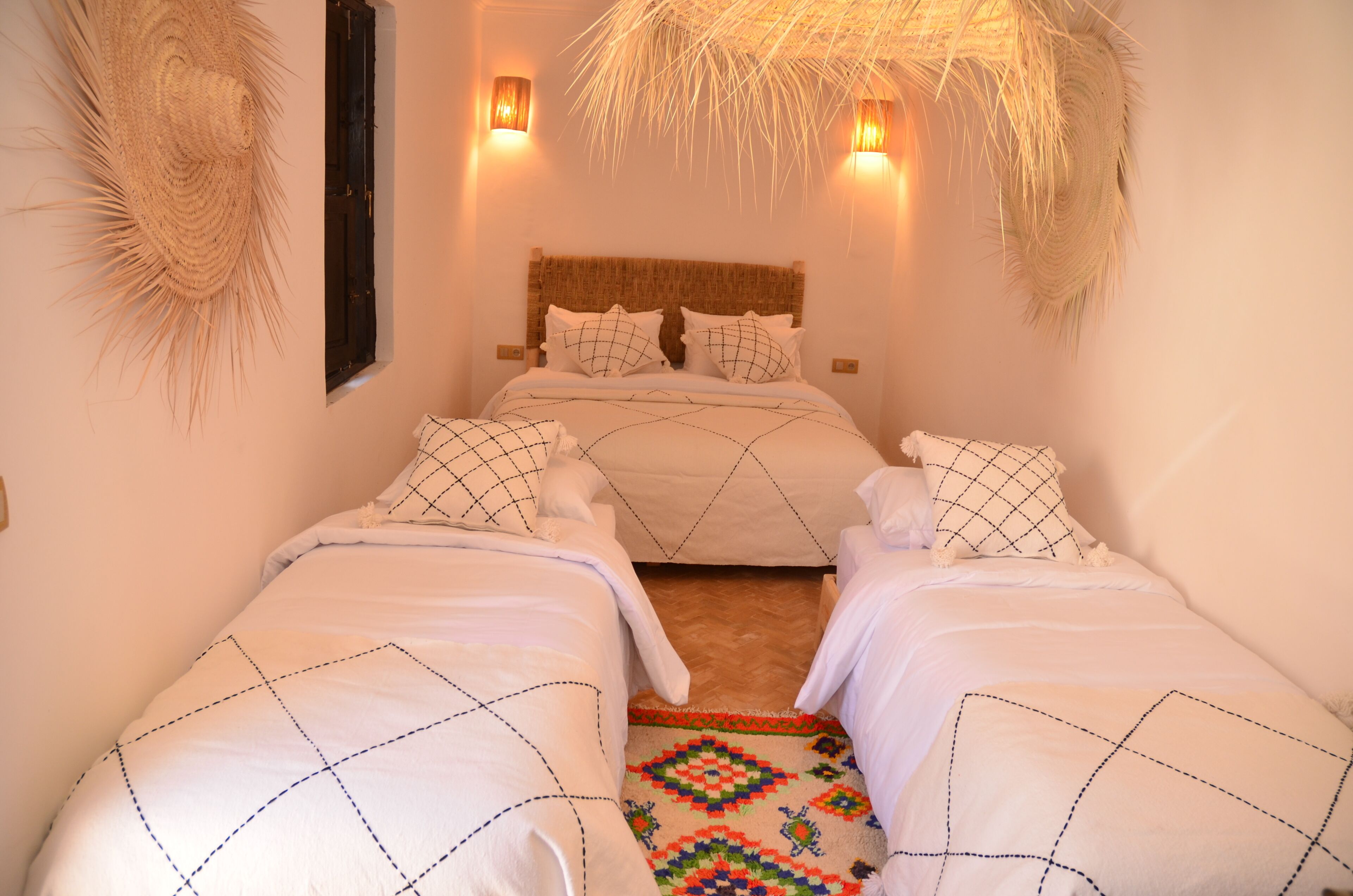 A room with two beds, a woven headboard, wall-mounted lights, and a patterned rug.