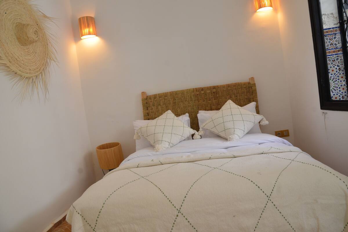 A bed with a woven headboard, two wall-mounted lights, a woven basket, and a textured wall decoration.