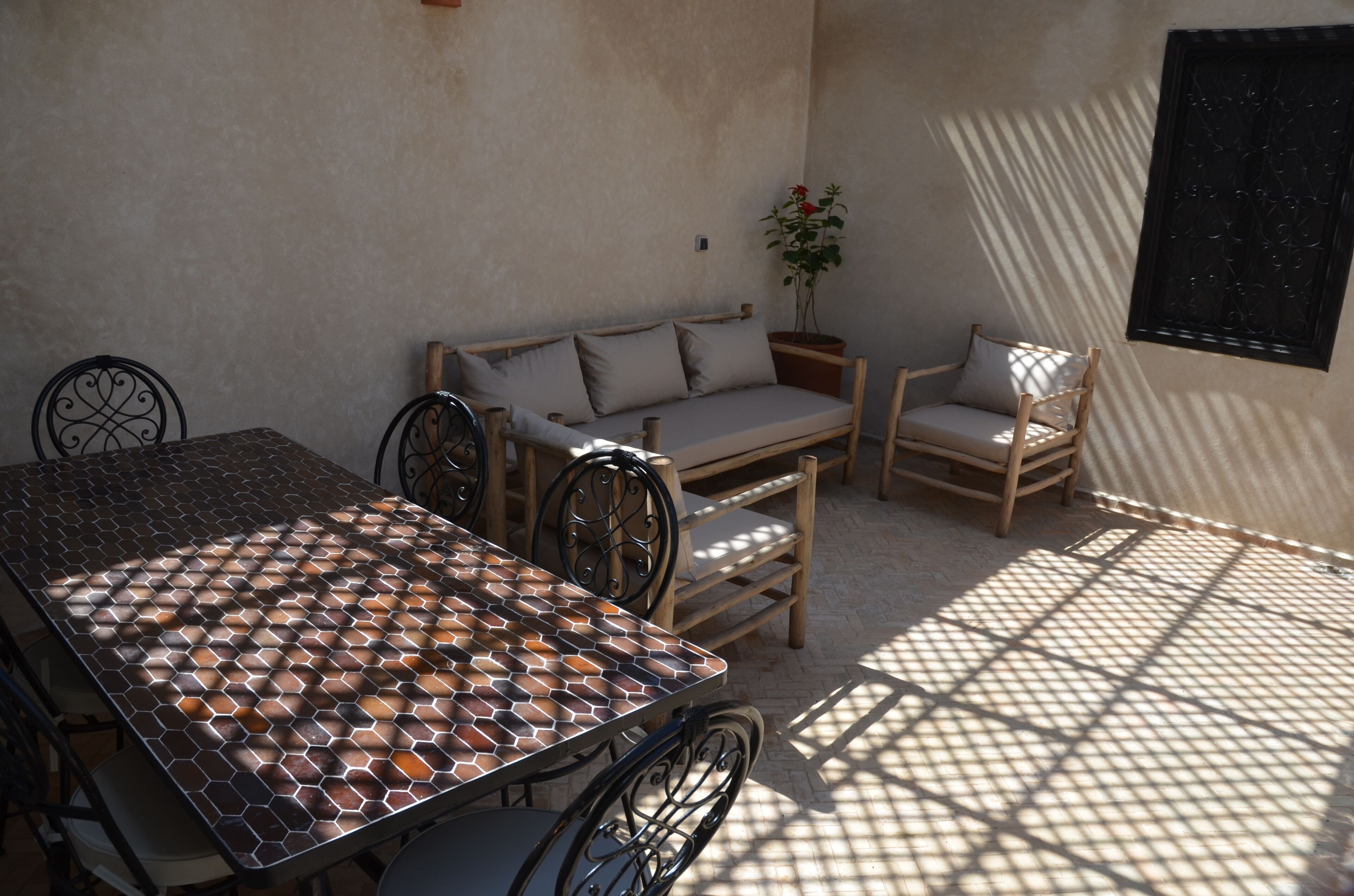 A sunlit patio with a patterned table, cushioned seating, and a potted plant.