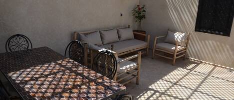 A sunlit patio with a patterned table, cushioned seating, and a potted plant.
