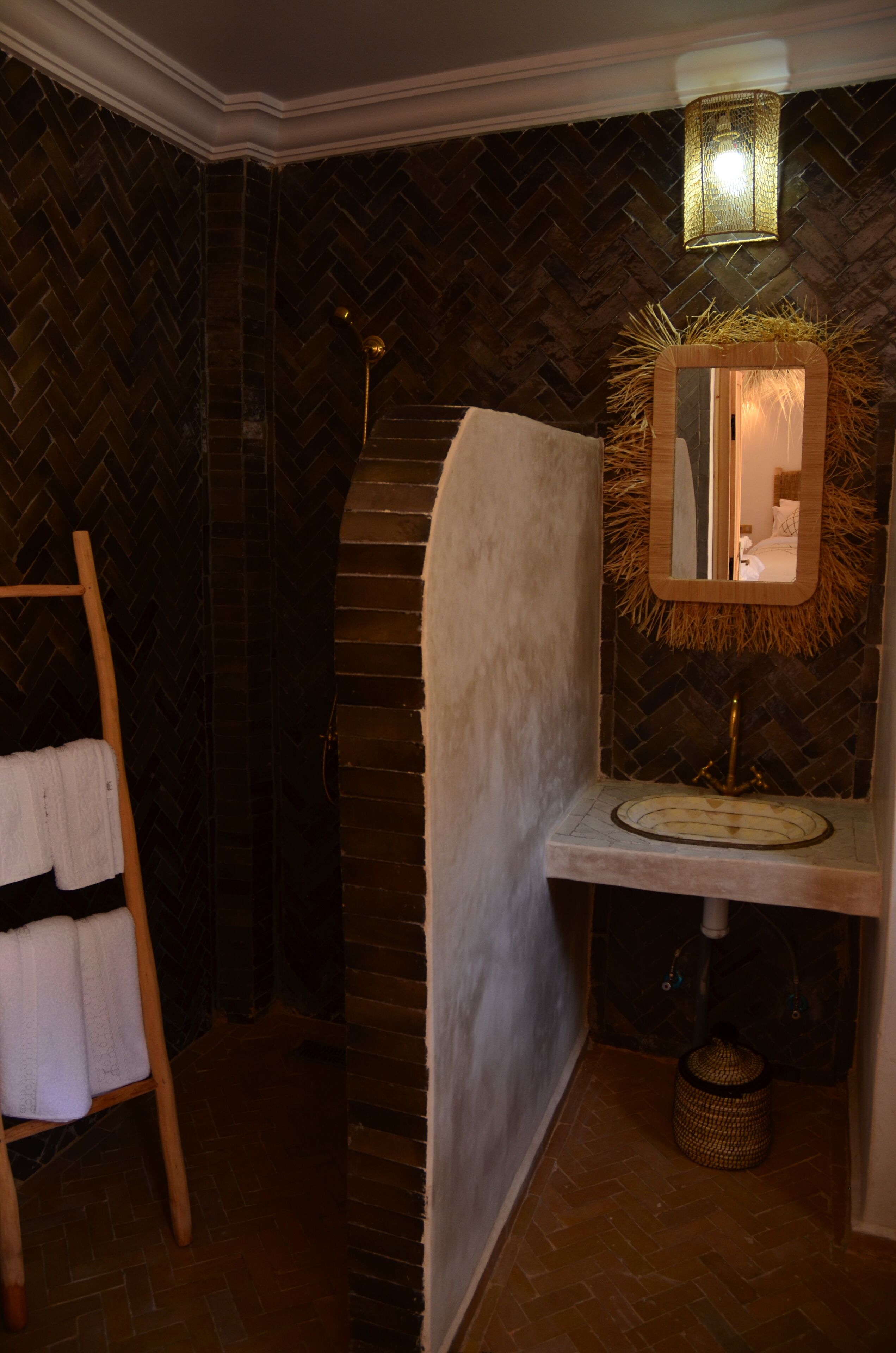 Une salle de bain avec un mur orné d’un motif en chevrons, un porte-serviettes en bois, un miroir décoré d’une couronne de paille et un lavabo avec un robinet doré.
