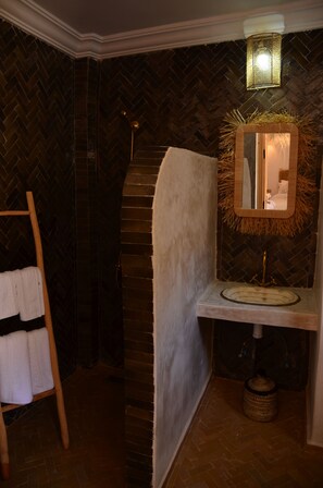 A bathroom with a herringbone-patterned wall, a wooden towel rack, a mirror with a straw wreath, and a sink with a gold faucet. - Riad Ayana (Marrakech)