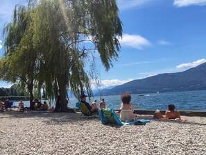A pebbled beach with people relaxing under a large tree, overlooking a lake with boats and mountains in the background. - A Moment of Peace by Interhome (Porto Valtravaglia)