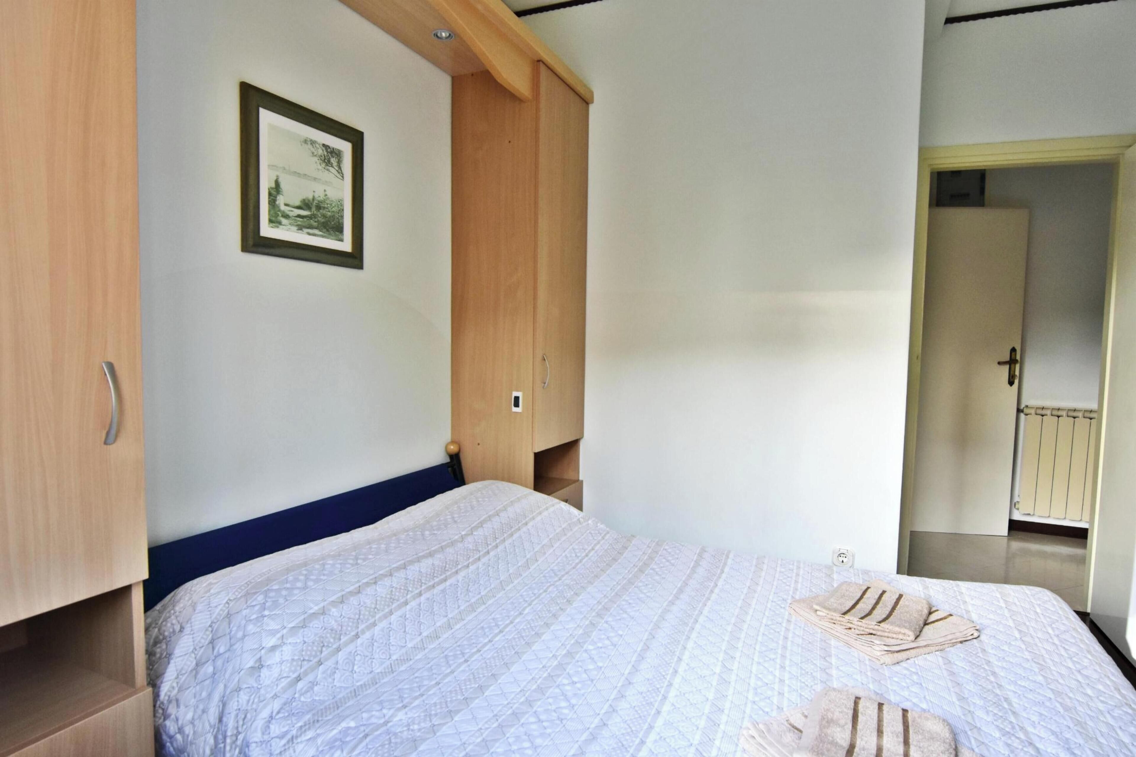 A bedroom with a bed, a wooden wardrobe, a framed picture on the wall, and a door leading to another room.