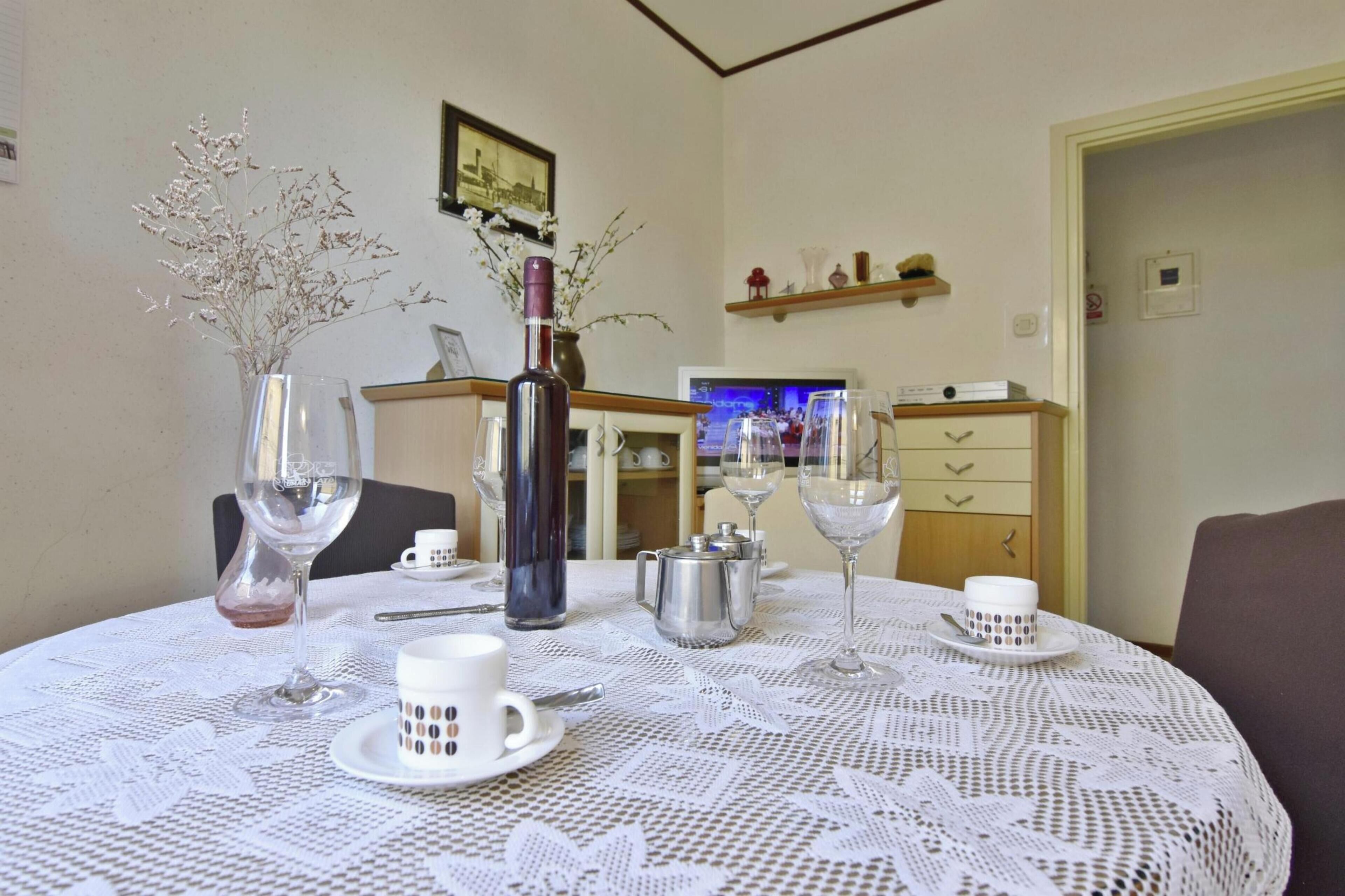 A dining table with a lace tablecloth, a vase with dried flowers, a wine bottle, and a coffee pot.