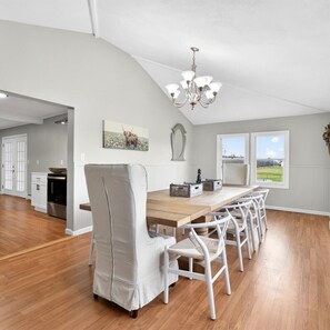 A spacious dining area with a long wooden table and white chairs, a chandelier, and a view of the outdoors through large windows. - Country Fields, Wine Country, Pool and More (Auburn)