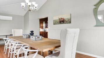 Un comedor con mesa de madera, sillas blancas, una araña y una cocina al fondo, además de un cuadro en la pared.