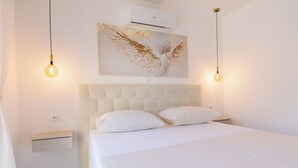 Un dormitorio con cabecera capitonada, ropa de cama blanca, dos mesitas de noche y un aire acondicionado de pared.