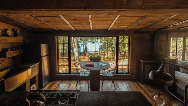A rustic cabin interior with a large window, wooden flooring, a round table, and a kitchen area.