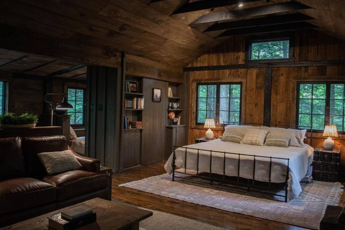 A rustic bedroom with a large bed, wooden walls, and a view of trees through windows.
