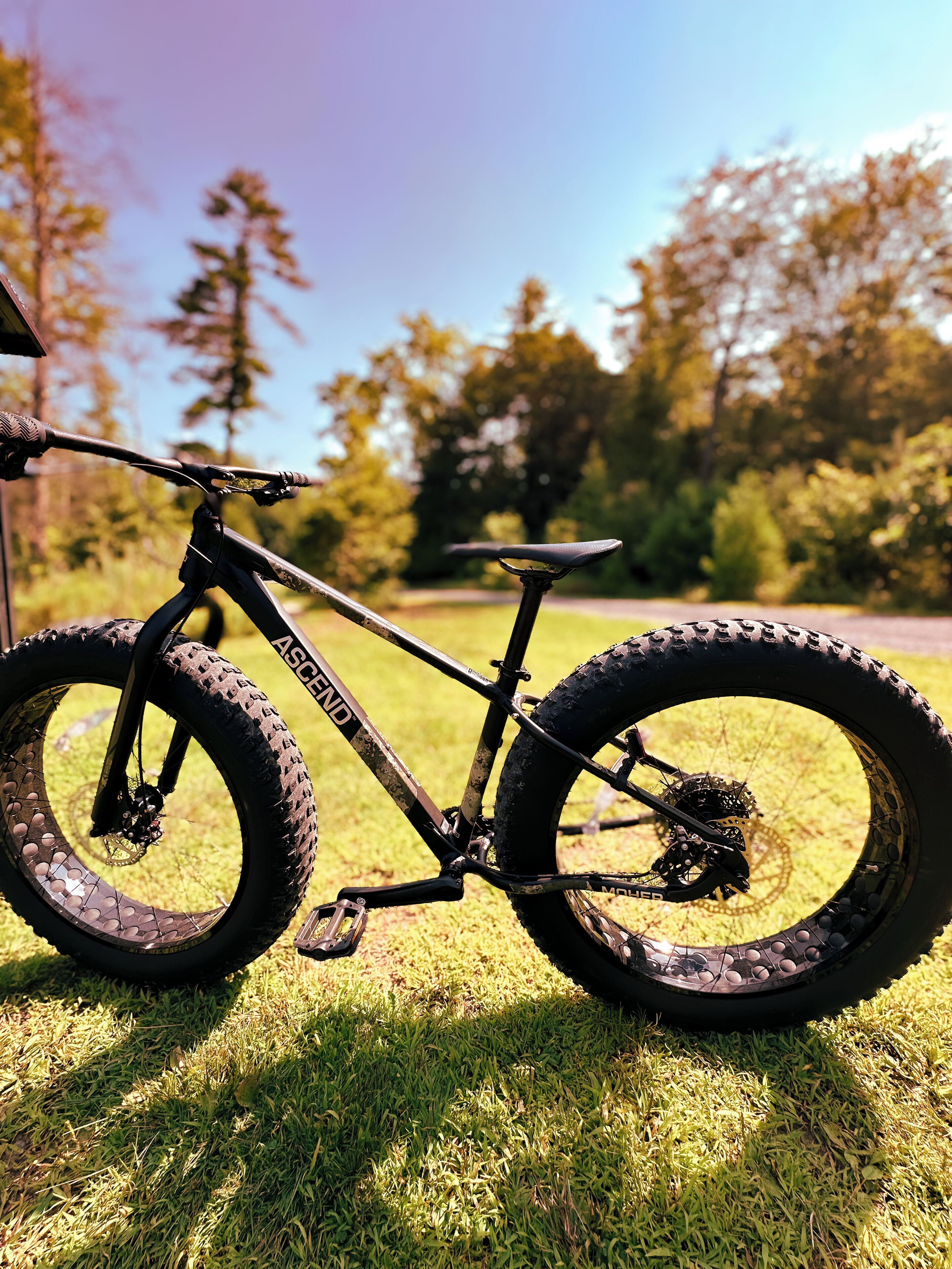 Een mountainbike met een zichtbaar merk op het frame, staand op gras met bomen op de achtergrond.