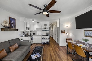 Een moderne keuken met witte kasten, een eethoek met een houten tafel en stoelen, een grijze bank en een plafondventilator.