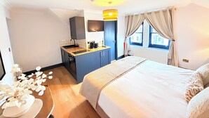 Hotellihuone, jossa on sänky, pieni keittokomero, ikkuna verhoilla ja näkymä rakennuksiin.