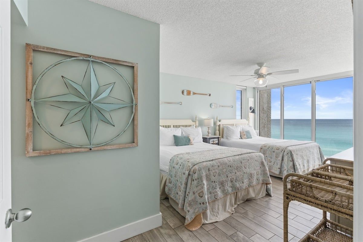 A bedroom with two beds, a large window with a view of the ocean, a ceiling fan, and a rustic wall decoration.