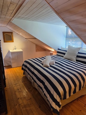 A bedroom with a sloped ceiling, a bed with striped bedding, a bedside table, and a mirror. - Loft Cottage - Knysna (Knysna)