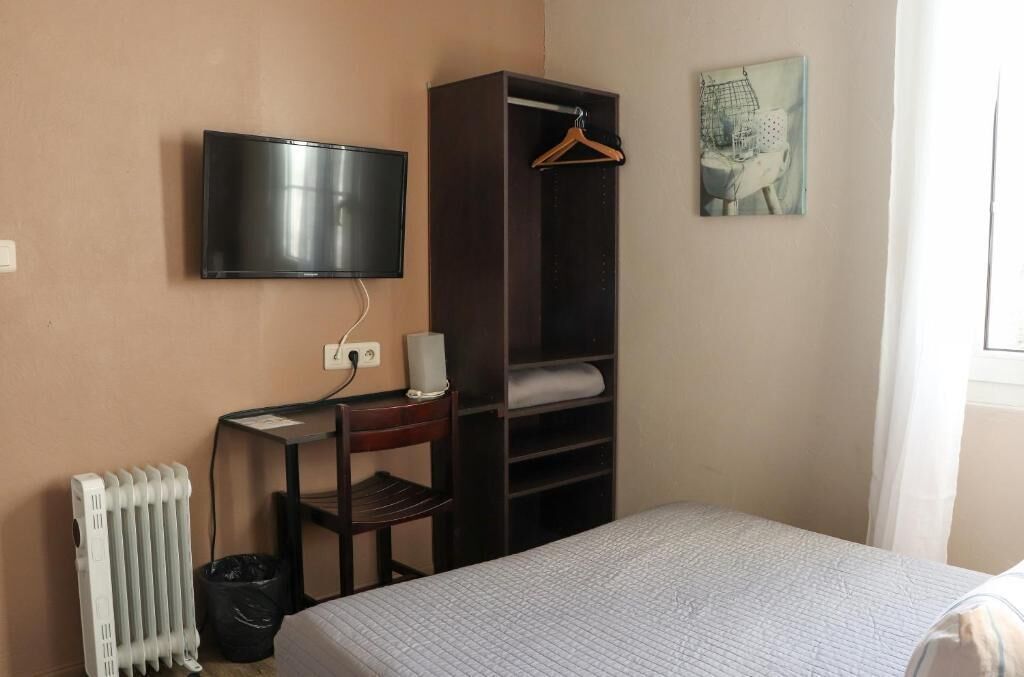 A hotel room with a bed, a TV mounted on the wall, a desk with a chair, a radiator, and a coat rack with a hanger.