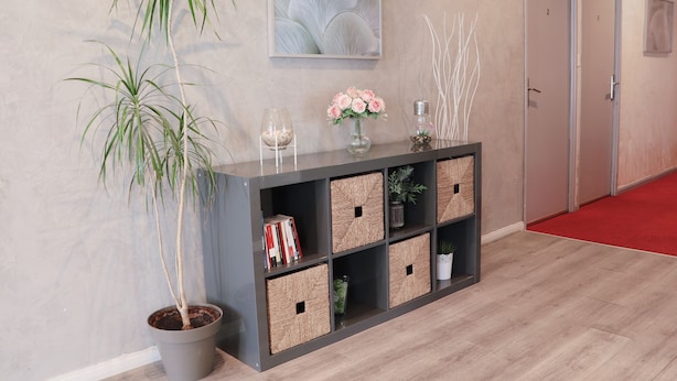 A reception area with a grey console, potted plants, a vase with flowers, and a framed artwork on the wall.