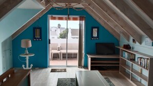 Ett rum med ett sluttande tak, en tv, en bokhylla och en utsikt genom en skjutdörr till ett utomhusområde med ett bord och stolar.