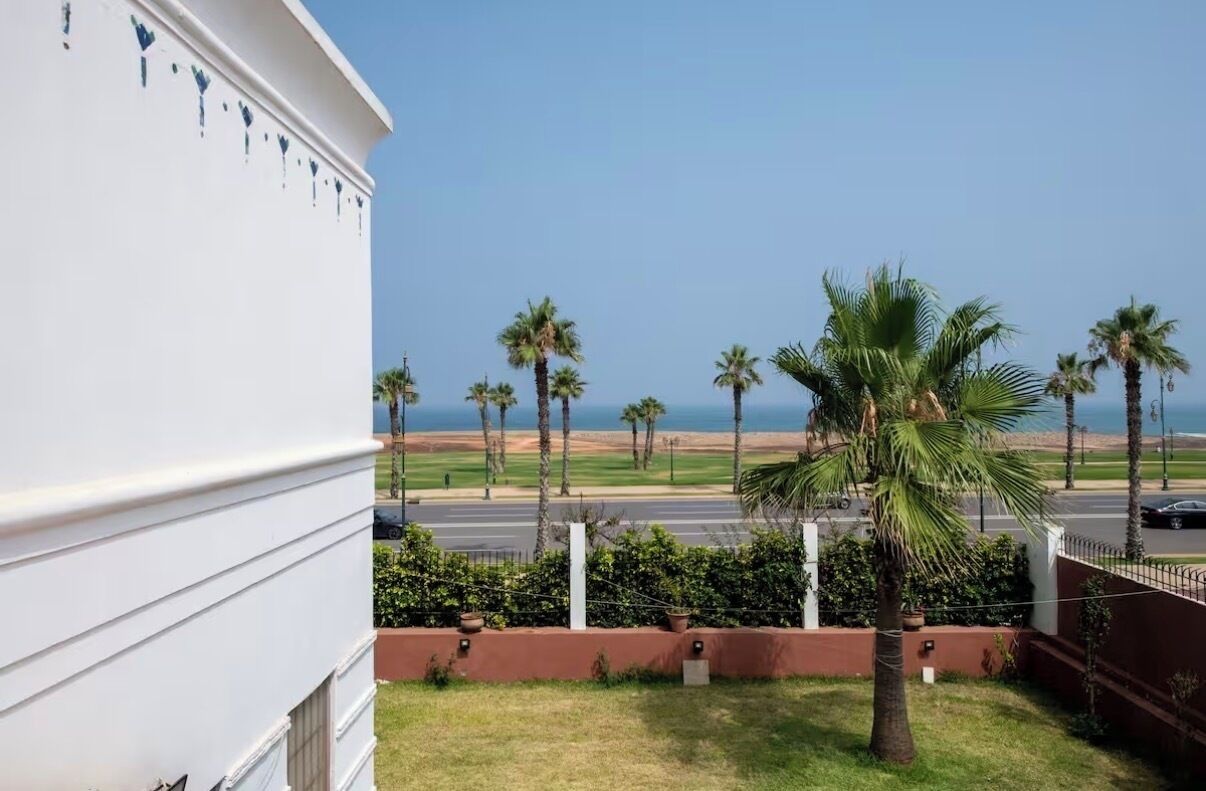 A building with a white facade, a palm tree in the foreground, and a view of the beach and ocean in the background.