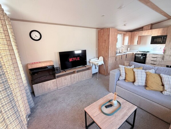A modern living room with a television, a sofa, a small table, a kitchen area with cabinets and appliances, and a clock on the wall.