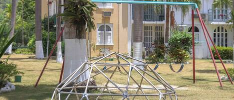 Un parque infantil con columpios y una estructura para escalar, en un área verde con palmeras y un edificio al fondo.
