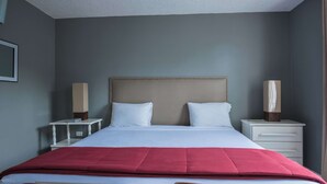 Un lit bien fait, avec une couverture rouge, deux oreillers blancs et une tête de lit beige. Il y a deux tables de chevet avec des lampes et une petite table de nuit avec une lampe.