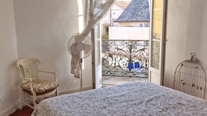 Quarto com cama, cadeira, ventilador e varanda com vista para edifícios.