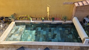 Piscina retangular com padrão de azulejos quadriculados, cercada por uma cerca de madeira, e duas espreguiçadeiras em primeiro plano.