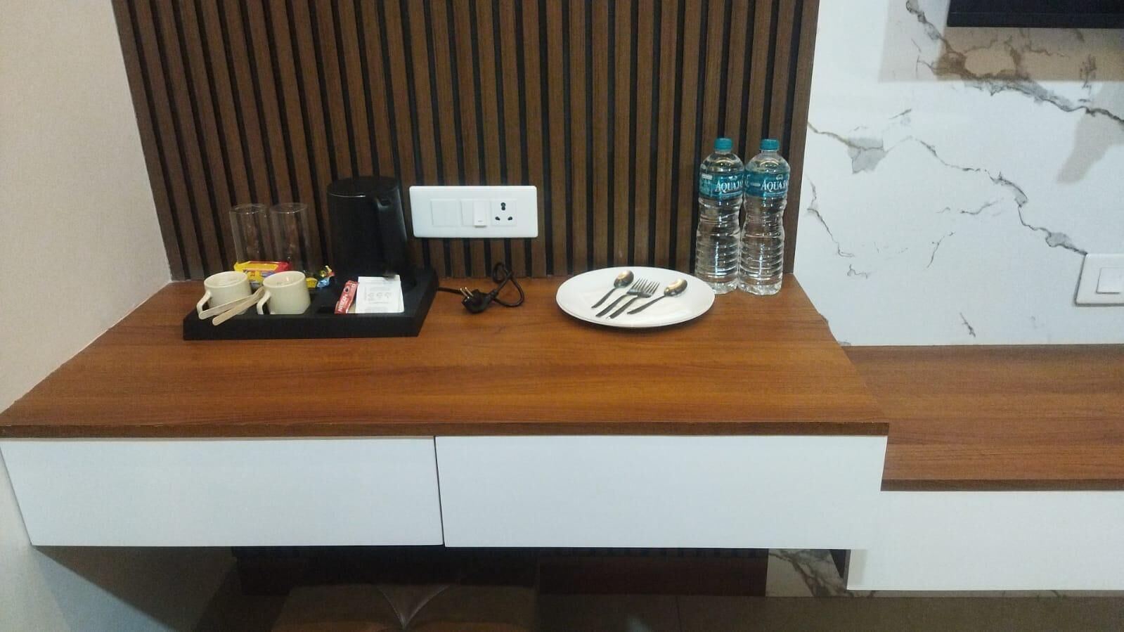 A wooden desk with a white drawer, a black coffee maker, two water bottles, a plate with cutlery, and a glass.