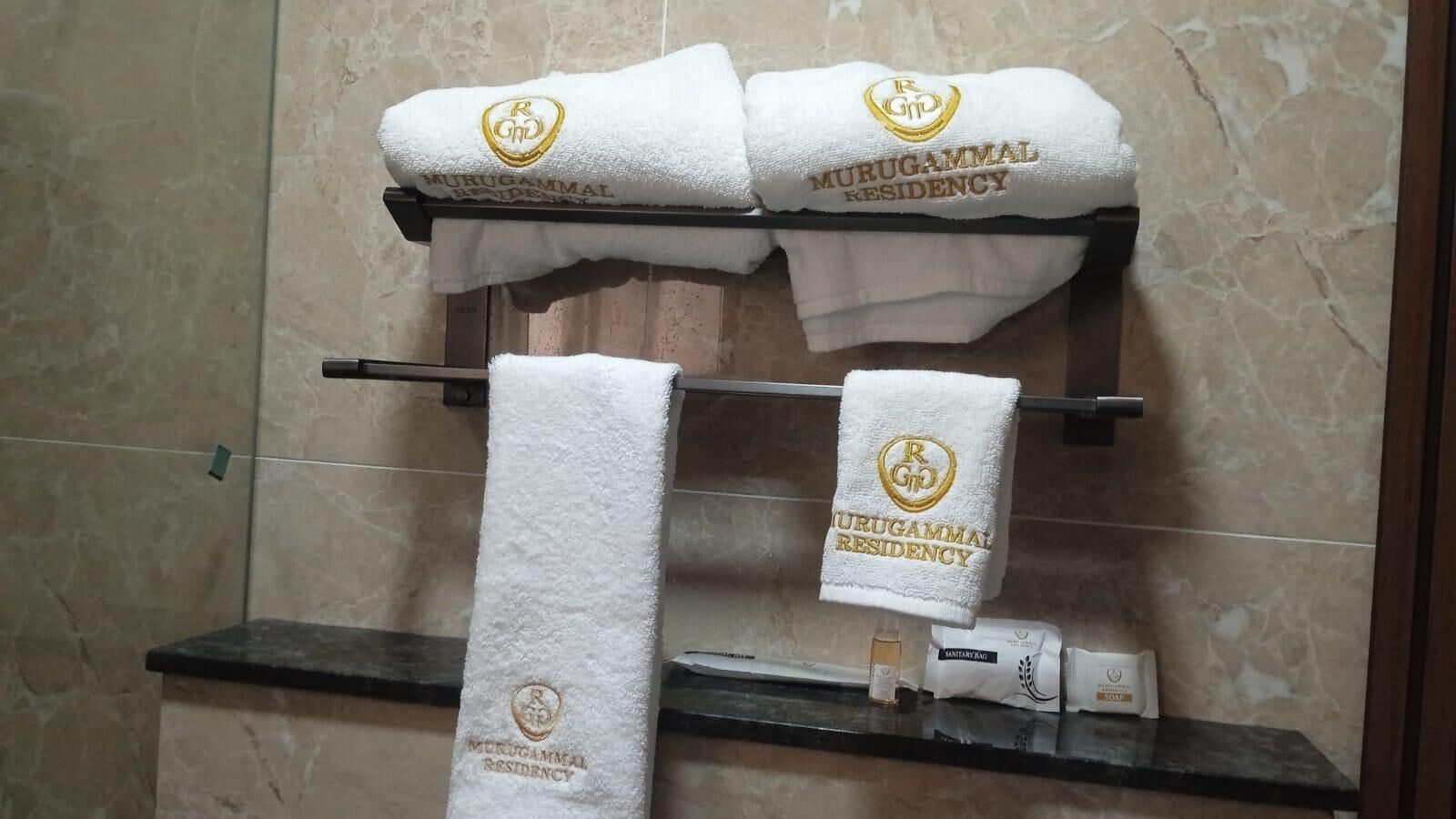 Un baño con dos toallas blancas colgadas en un perchero, cada una con un emblema dorado y el texto 'MURUGAMMAL RESIDENCY'.