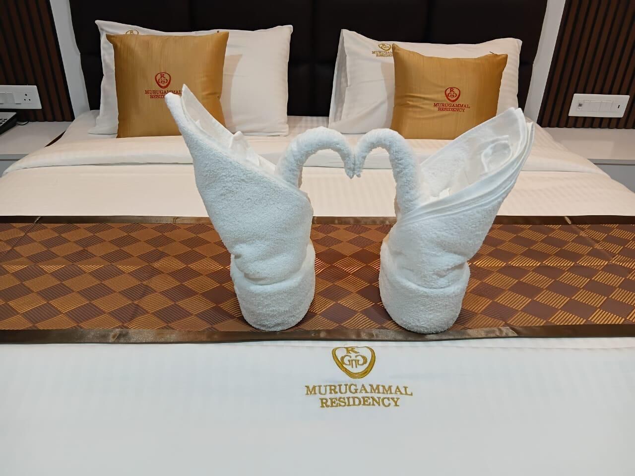 A hotel bed with a checkered bedspread, two towels arranged in a heart shape, and a sign indicating the hotel name.