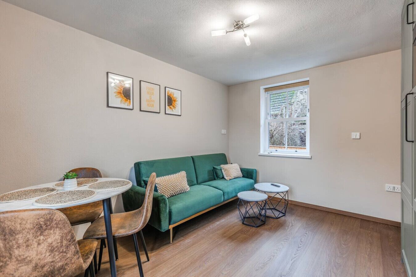 Une pièce avec un canapé vert, une table de salle à manger ronde, deux petites tables d’appoint, un ventilateur de plafond et une fenêtre donnant sur un beau paysage.