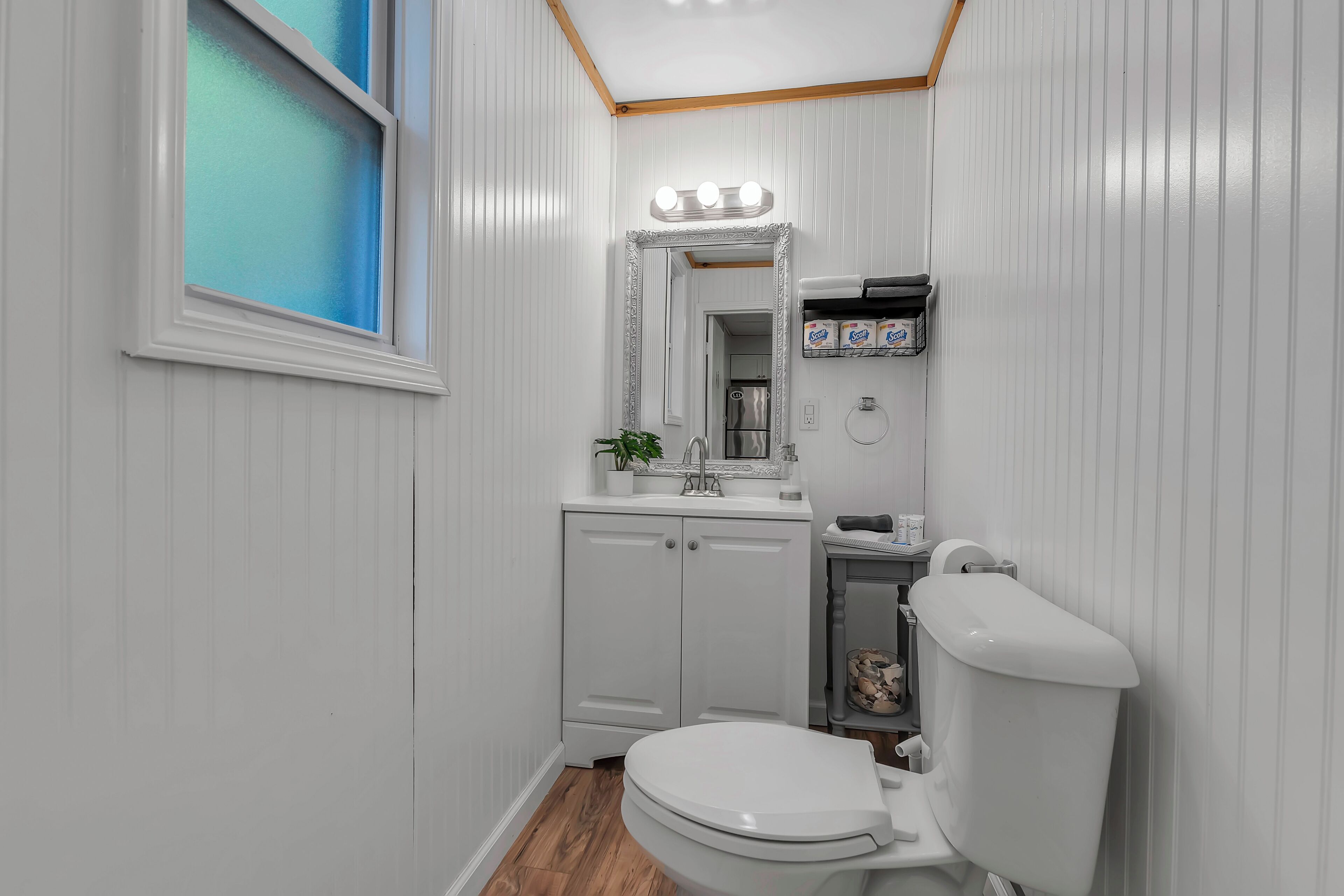 Une salle de bain avec des toilettes blanches, un lavabo surmonté d’un miroir et une fenêtre à vitre dépolie.