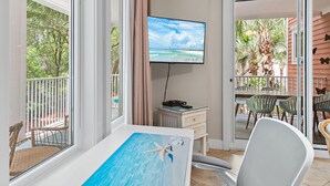 A room with a TV, a painting of a swimming starfish, a dining area with chairs, and a view of a balcony with outdoor furniture. - 🦀Lagoon Pool | Hot Tub | Walk to Baytowne Wharf | Market Street Inn 232/234🐚 (Miramar Beach)