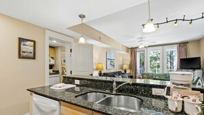 Cozinha com bancadas de granito, pias de aço inoxidável e lava-louças. Há uma área de refeições com mesa e cadeiras, e uma TV fixada na parede.