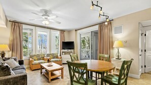Uma sala de estar ampla com ventilador de teto, televisão, mesa de jantar com cadeiras e janelas grandes com cortinas.