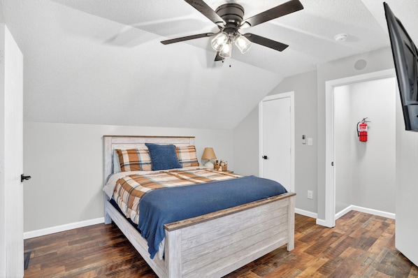 A bedroom with a bed, a ceiling fan, a nightstand, a lamp, and a fire extinguisher.