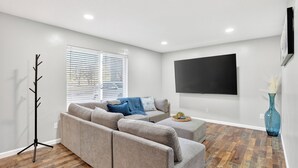 A modern living room with a sectional sofa, a flat-screen TV, a floor lamp, and a large window with blinds. - Newly Remodeled Condo in the Heart of Springfield (Springfield)