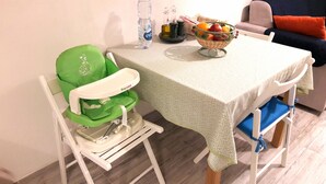 Et værelse med et spisebord, en grøn babystol, en kurv med frugt og en vandflaske.