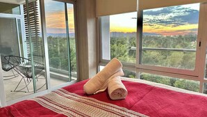 Ein Hotelzimmer mit Bett, roter Decke, gerolltem Handtuch und einem Balkon mit Blick auf Sonnenuntergang und Stadtlandschaft.