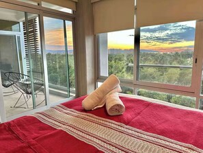 Ein Hotelzimmer mit Bett, roter Decke, gerolltem Handtuch und einem Balkon mit Blick auf Sonnenuntergang und Stadtlandschaft.