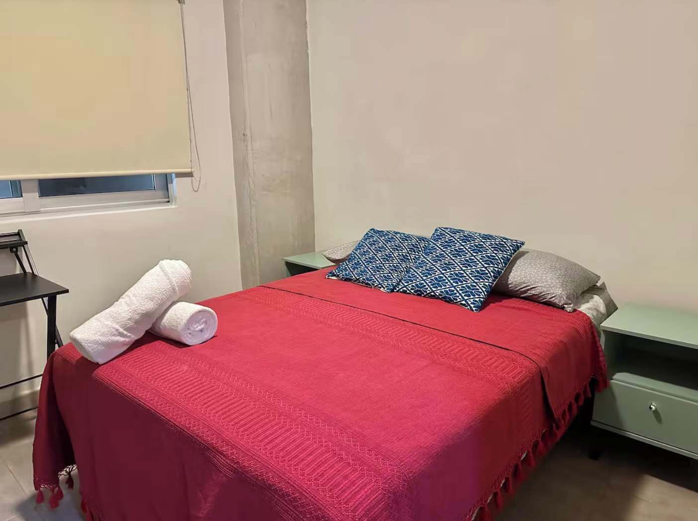 A single bed with a red bedspread, two pillows, a white towel, a green nightstand, and a small table with a chair.