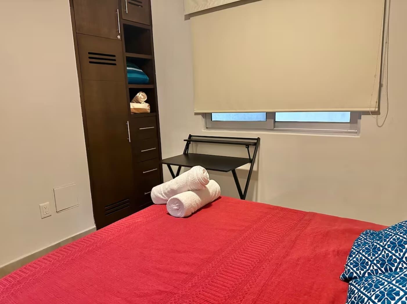 A bedroom with a red bedspread, a small table, a window with blinds, and a wardrobe with shelves.