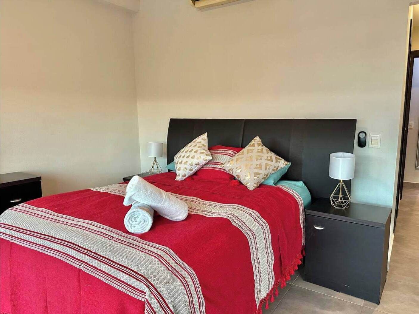 A neatly made bed with a red blanket, two patterned pillows, and a rolled towel. There are two bedside tables with lamps, one on each side of the bed.