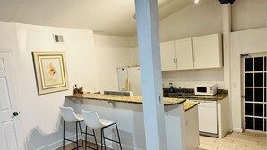 Una cocina con armarios blancos, microondas y lavabo. Hay dos taburetes blancos y un cuadro en la pared.