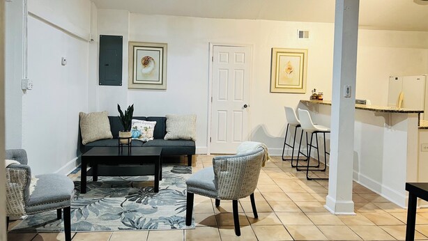 Una sala de estar con sofá, sillas, una mesa de centro, un ventilador de techo y una cocina con refrigerador y taburetes de bar.